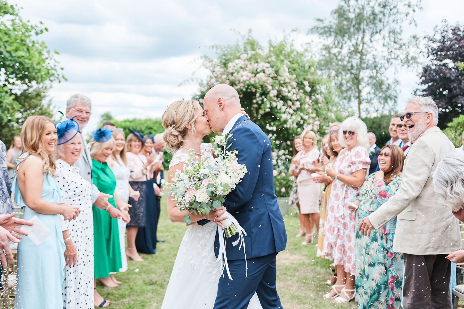 bride and groom kiss as guests smile and throw confetti. by award winning norfolk wedding photographer Faye Amare