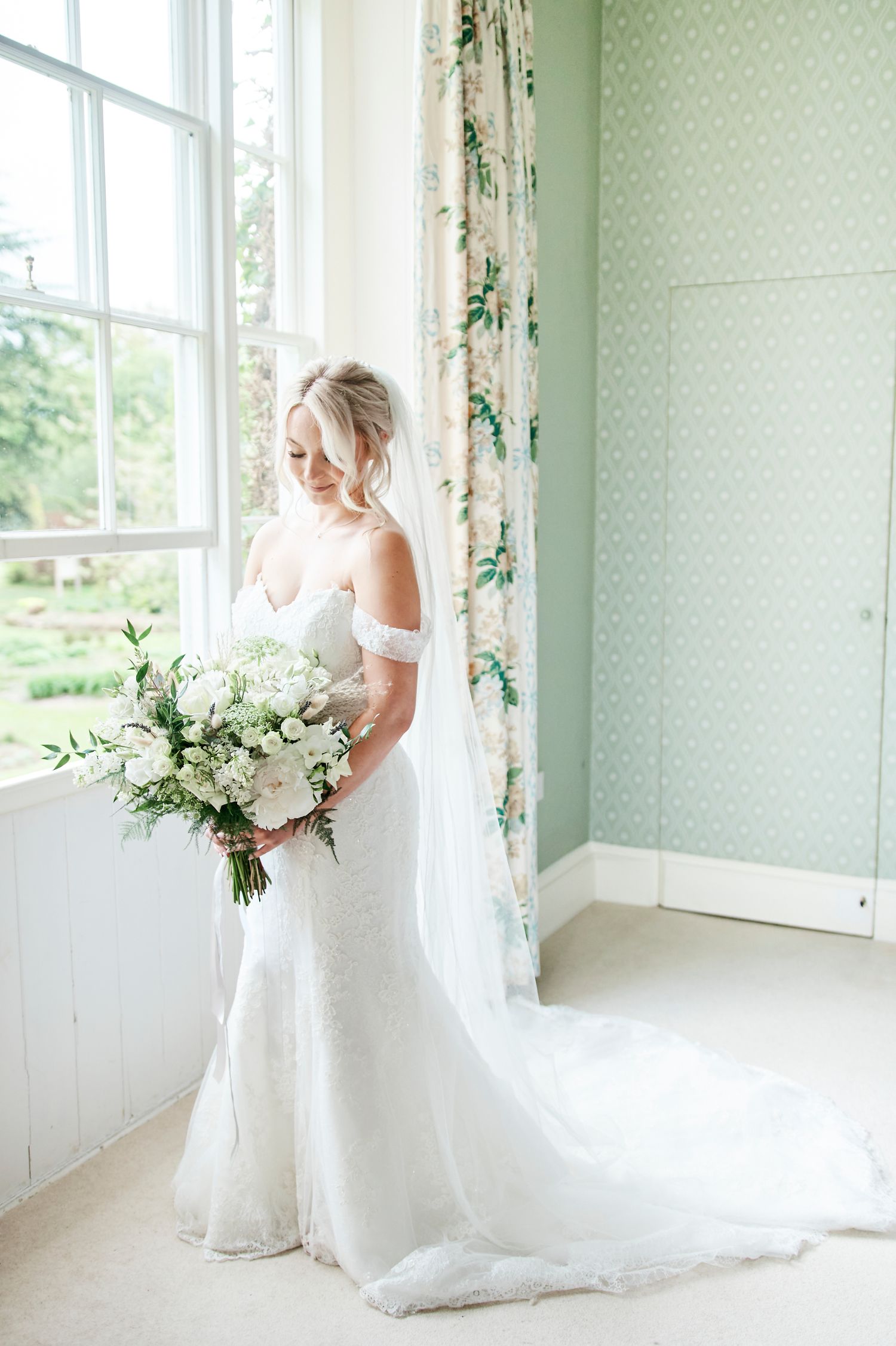 stunning bride looks down at her bouquet in her dress at luxury wedding venue bressingham Hall, Suffolk. by Faye Amare