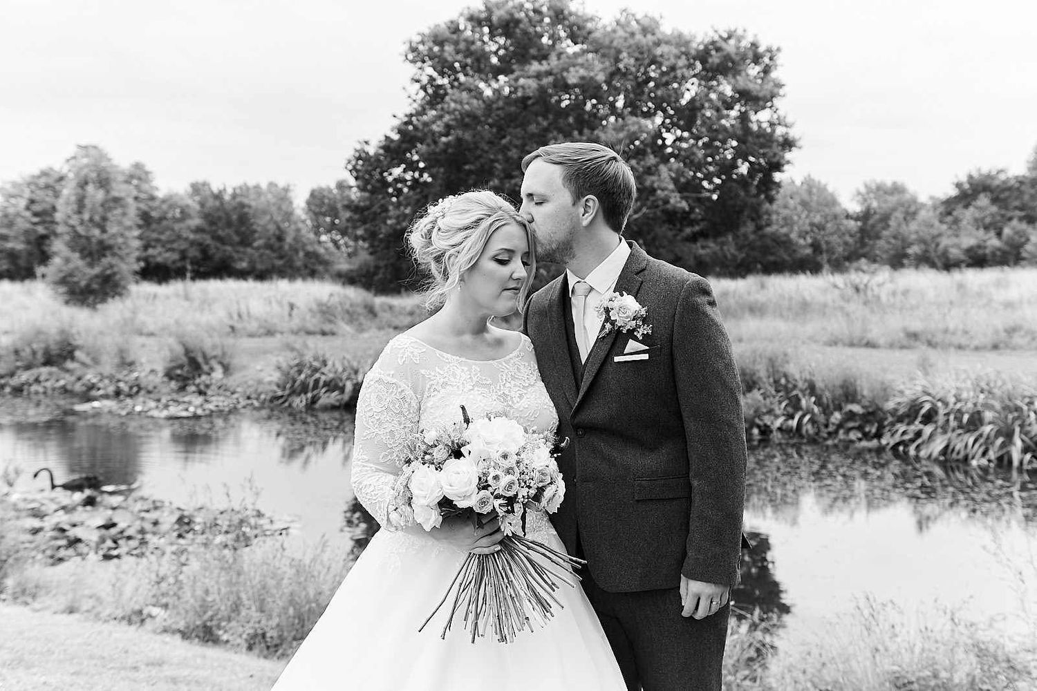 norfolk wedding photographer groom kisses his bride on the temple as they stand in front of a beautiful norfolk lake